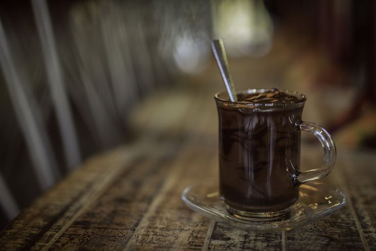 A warm chocolate beverage in a glass mug with saucer on a rustic wooden table, featuring a blurred bokeh background.